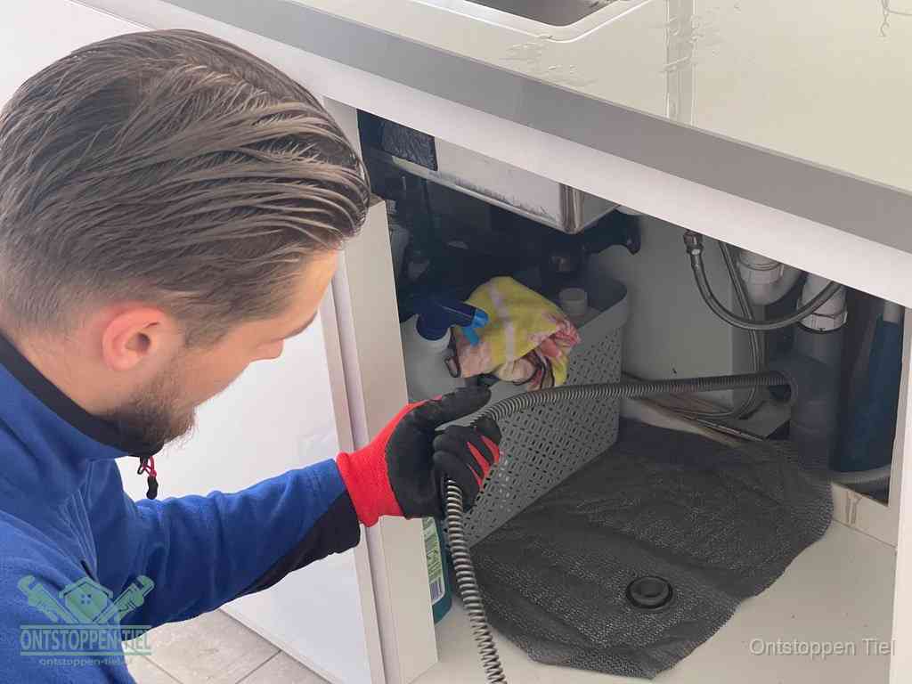Een monteur voert een flexibele ontstoppingsveer in onder een keukenkastje om een verstopping voorbij de sifon te bereiken in Tiel