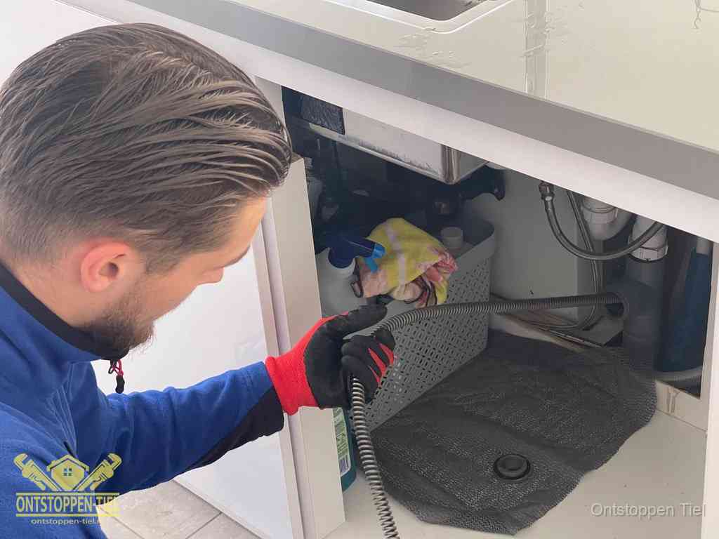 Een monteur voert een flexibele ontstoppingsveer in onder het keukenkastje, langs de sifon, om de gootsteen te ontstoppen in Tiel