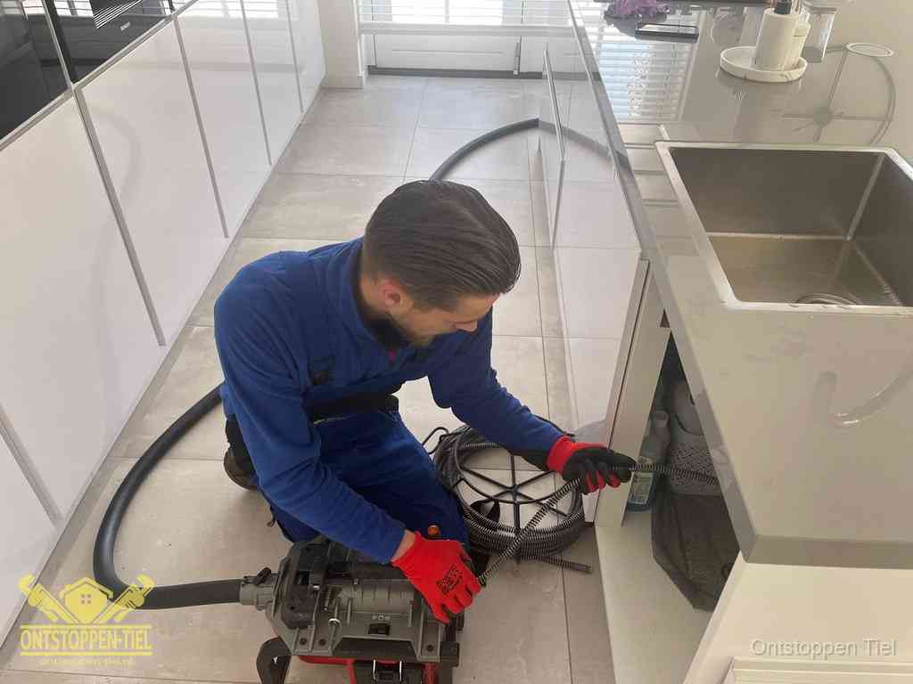 Een monteur ontstopt de keukenafvoer onder de spoelbak met een elektrische veermachine in Tiel