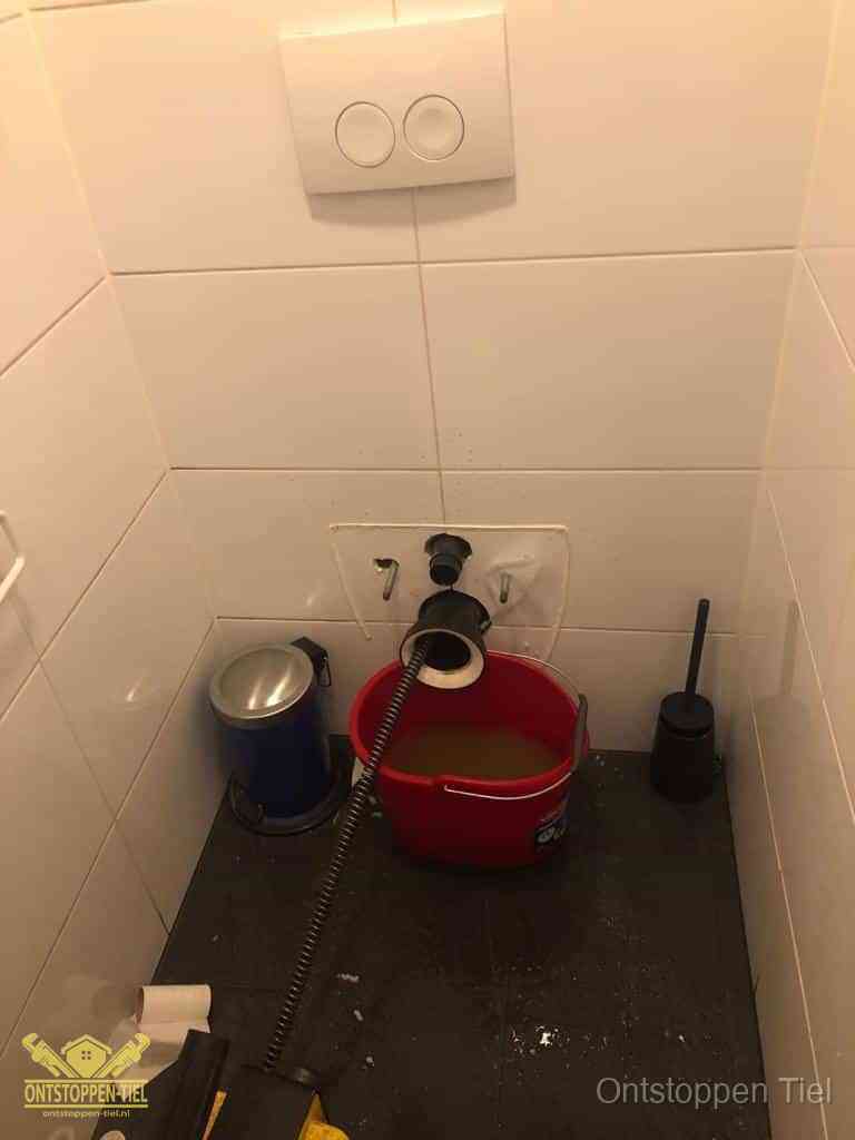 Een ontstopte wand-wc met verwijderde toiletpot waar een ontstoppingsveer in de wanduitgang verdwijnt, het achterhalen waar de verstopping zit in Tiel