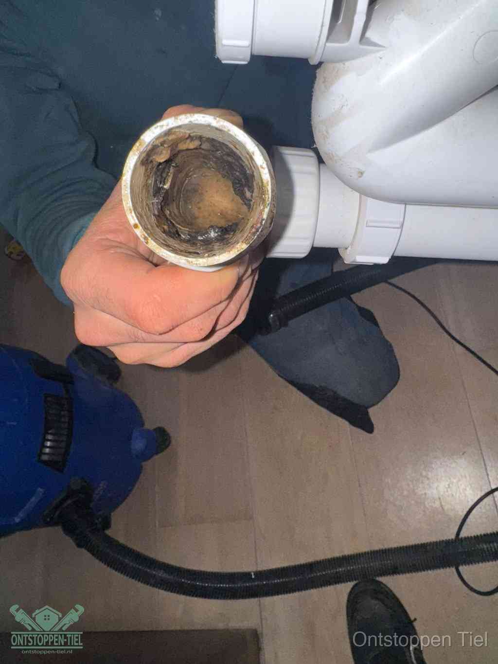 Een hand houdt een losgekoppeld PVC-afvoerbuisstuk vast dat van binnen volledig is dichtgeslibd met vet, vuil en aanslag, de meest voorkomende oorzaak van een verstopte afvoer in Tiel