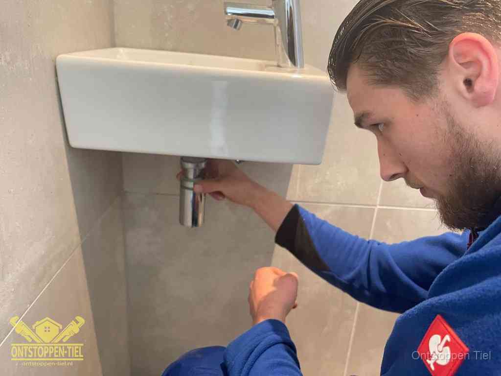Een monteur draait met de hand de zwanenhalssifon onder een fonteinwasbak in de toiletruimte los om de bron van rioollucht in Tiel te vinden