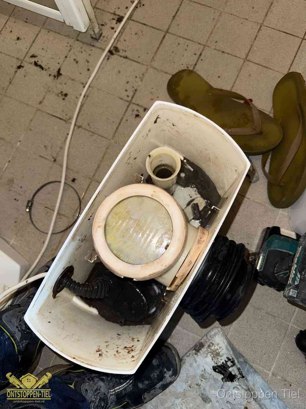 Een gedemonteerde toiletpot op de vloer met ontstoppingsveer en gereedschap ernaast, het verwijderen van een vastzittend voorwerp uit de wc in Tiel