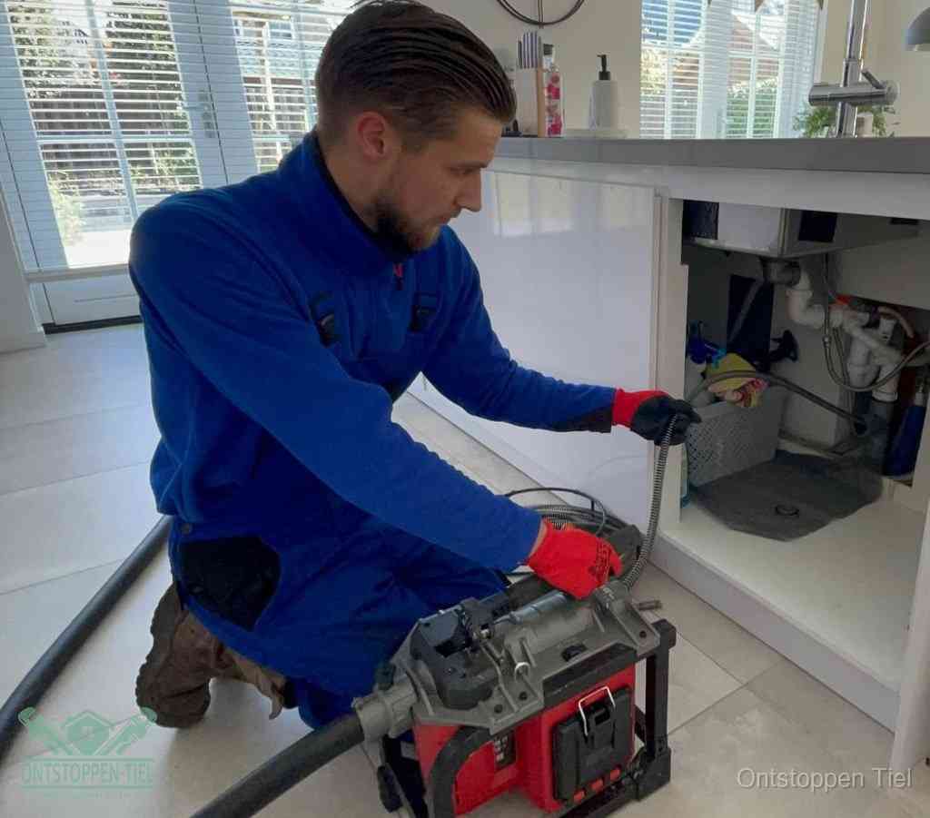 Een monteur met rode handschoenen knielt in een keuken en voert een elektrische ontstoppingsveer in tijdens het professioneel afvoer ontstoppen in Tiel